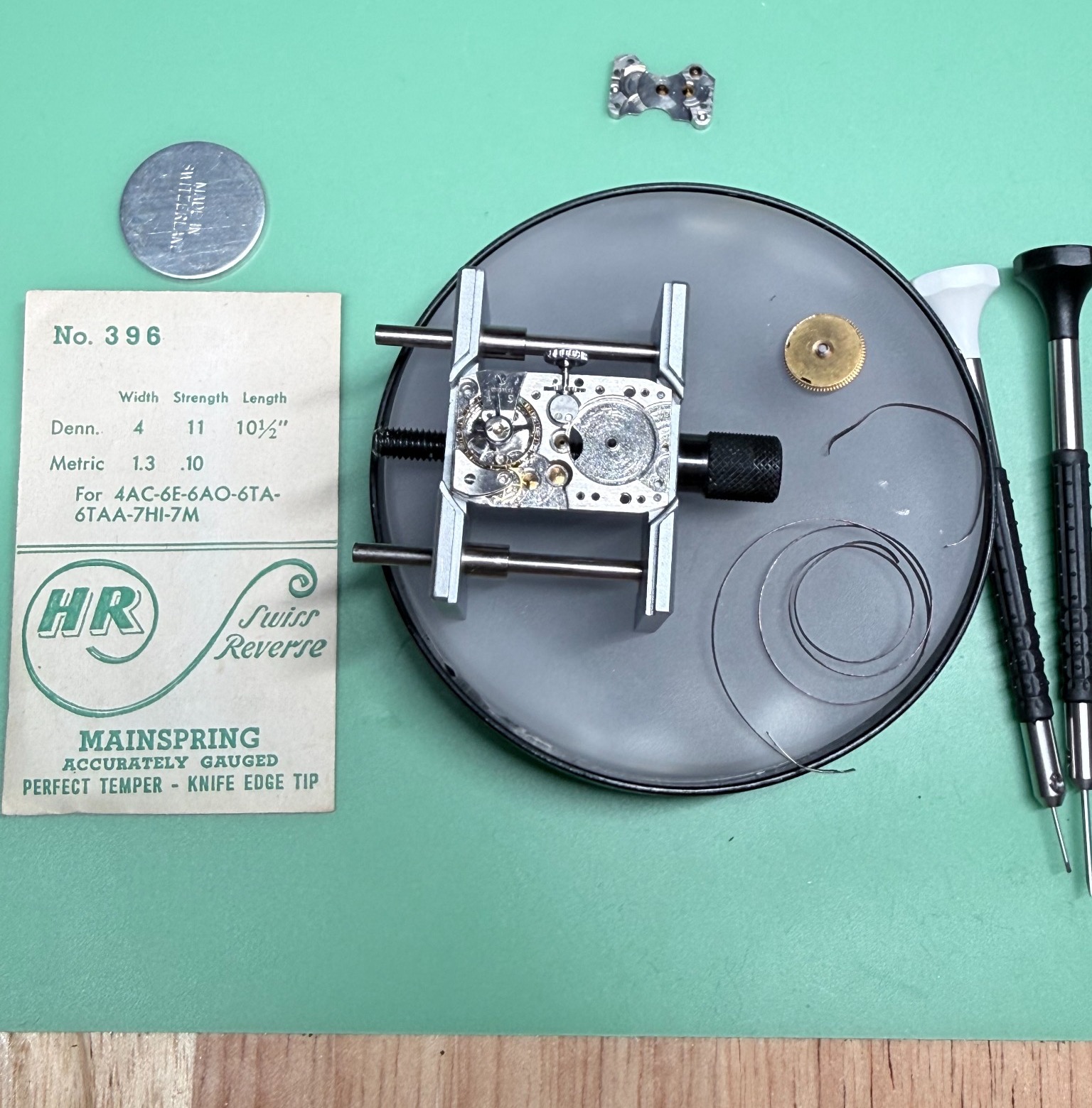 Vintage Elgin watch movement on a watchmaker’s bench beside a replacement mainspring and barrel components.